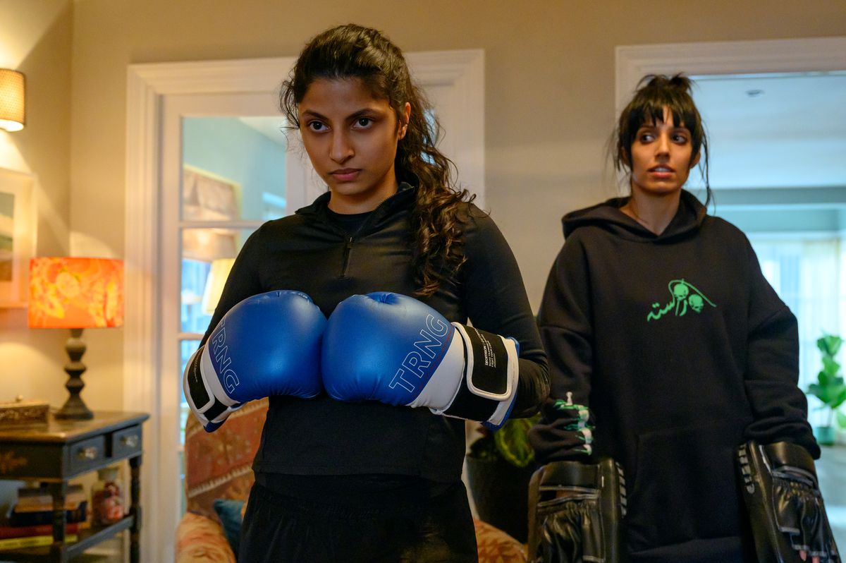 Ria, a British-Pakistani teenager, wears a black shirt and boxing gloves, which she presses together and looks determined.  Behind her is her sister Lena, who wears a black hoodie and holds two strikes that Ria can hit.  They are currently down, however, when it comes to Ria.